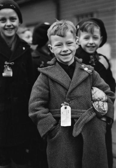 Dutch Child Refugees: Arrival In Britain At Tilbury, Essex, England, UK, 1945, A small Dutch boy smiles for the camera upon arrival at Tilbury in Essex. He is carrying a small paper parcel under his arm, which contains all his luggage. He, and the other children, (some of whom can be seen behind him) all have labels pinned to their coats which bear their names, home address and destination, 11 March 1945. (Photo by Ministry of Information Photo Division Photographer/ IWM via Getty Images)